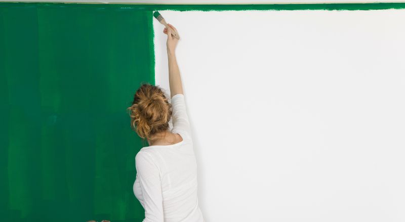 femme avec un pinceau en train de repeindre un mur blanc en vert traces de rouleau visibles avec absorption inegale de la peinture