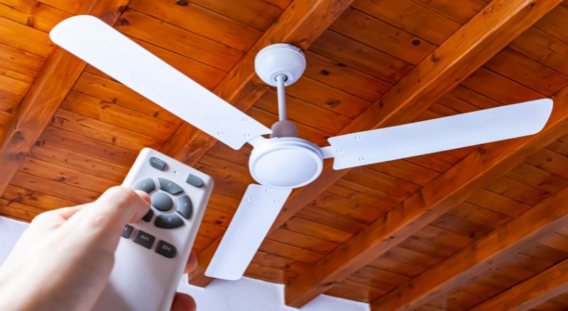 un homme utilise une telecommande pour allumer un ventilateur de plafond blanc monte dans une maison avec des plafonds en bois